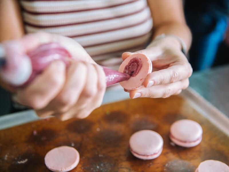 Close up of hands piping raspberry filling into a pink maracon
